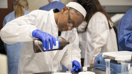 A student wearing protective gear pours a liquid from a large brown container into a tall clear container