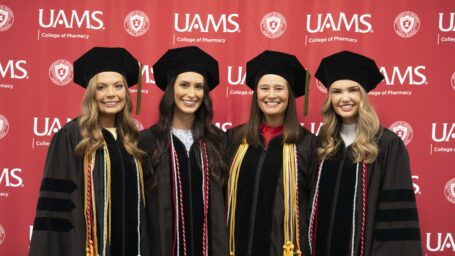 Four women in doctoral regalia smile and pose in front of a red UAMS sign.