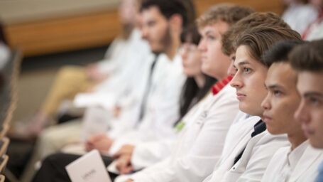 A row of students in white coats look towards a presentation (out of shot)