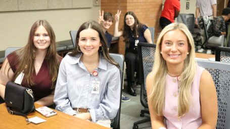 Three students sitting at a table, smiling at the camera, with more students in a classroom behind them.