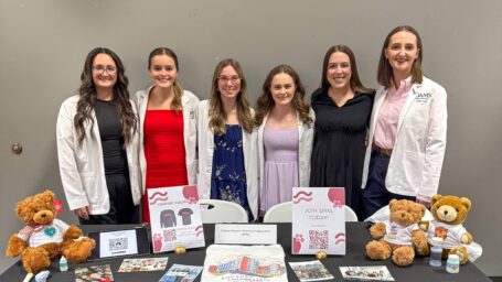Six students in white coats pose behind a table with information about SPPA, including photos, teddy bears, and QRs