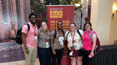 Five students pose in front of a Phi Delta Chi banner