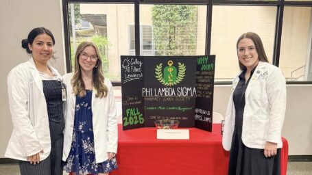 Three students pose alongside a booth with a poster promoting Phi Lambda Sigma