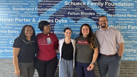 Five students pose in front of a blue wall with small white text