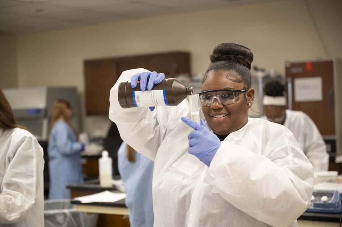 A student in a protective gown and latex gloves smiles and she stares closely as she pours liquid from a large brown bottle to a clear tall container