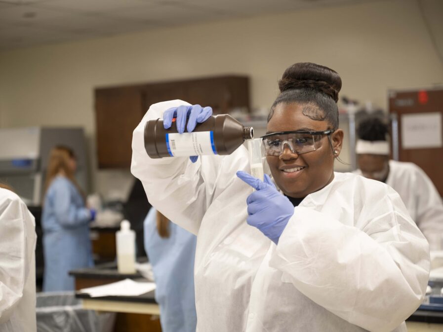 A student in a protective gown and latex gloves smiles and she stares closely as she pours liquid from a large brown bottle to a clear tall container