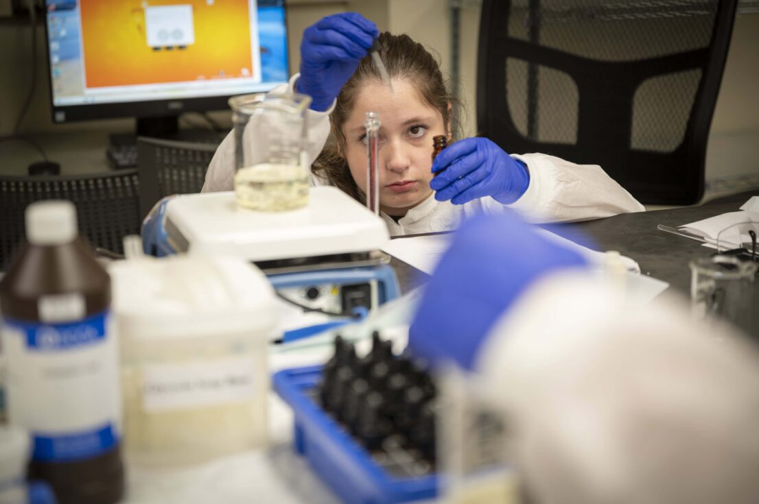 A student wearing a protective gown and latex gloves squats down as she gets a syringe of liquid from a small brown bottle into a tall clear container