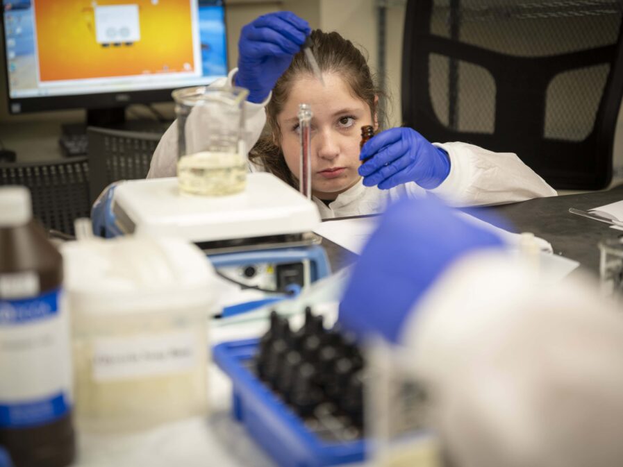 A student wearing a protective gown and latex gloves squats down as she gets a syringe of liquid from a small brown bottle into a tall clear container
