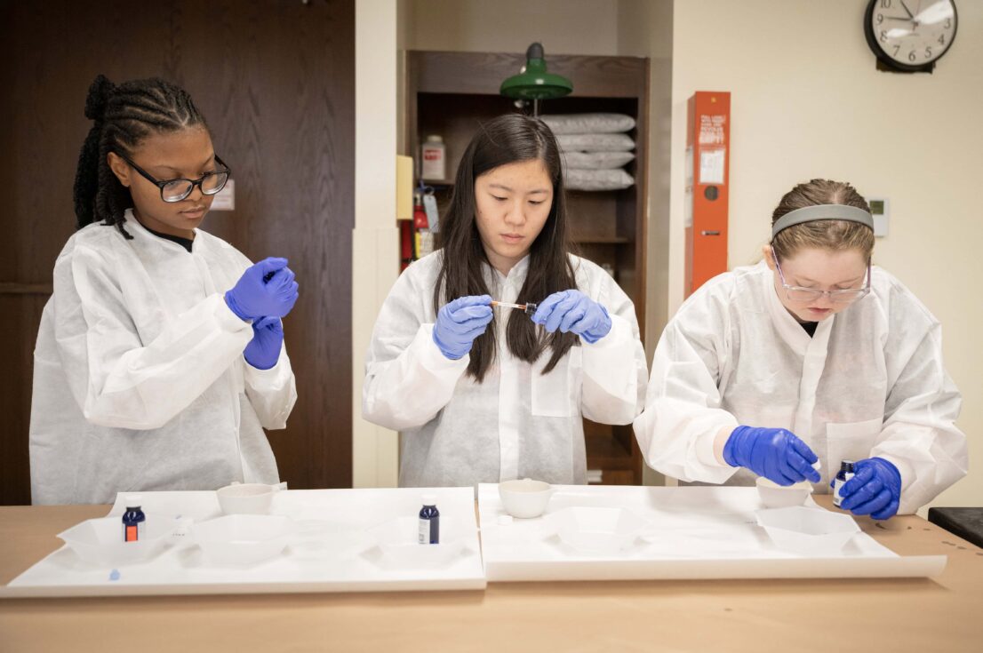 Three students, each in protective gowns and latex gloves, each look at their individual vials and assess the substance