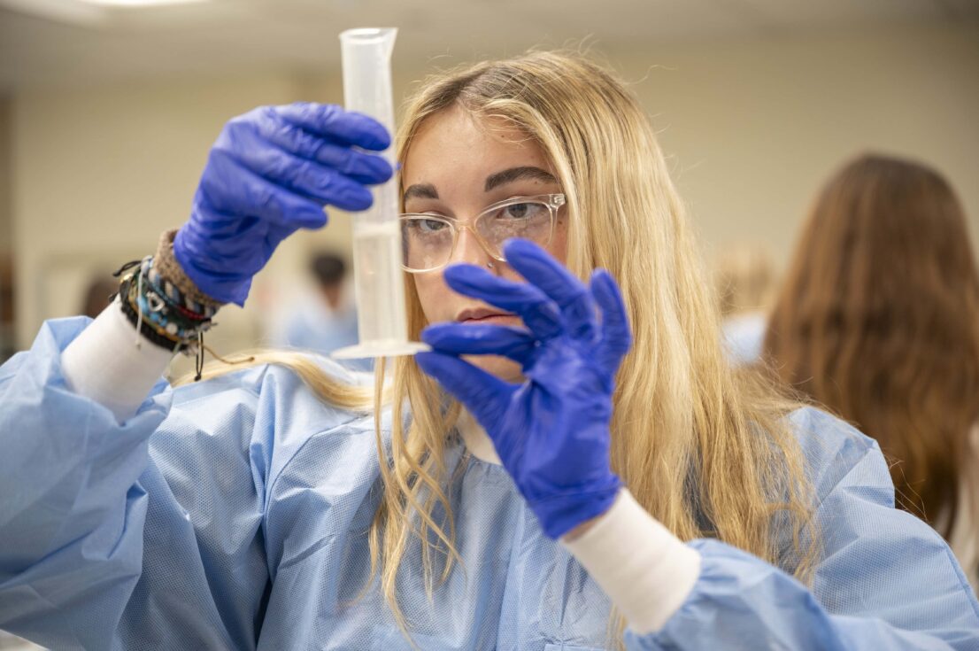 A student in a protective gown and latex gloves holds a clear container of liquid close to measure the volume.