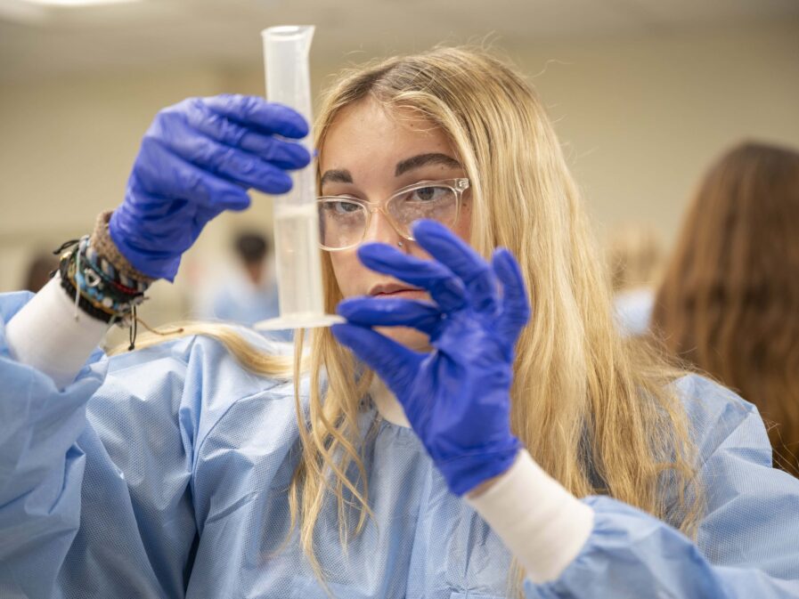 A student in a protective gown and latex gloves holds a clear container of liquid close to measure the volume.
