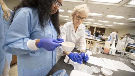 A professor in a white coat and gloves leans in to assist a student in a blue gown and blue gloes
