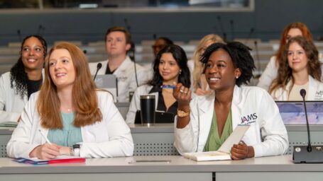 Two students sitting at a table with rows of students behind them engaging in a classroom discussion