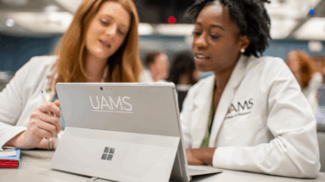 Two students, talking and wearing white coats with the UAMS logo, look at a tablet with a UAMS logo on the back.