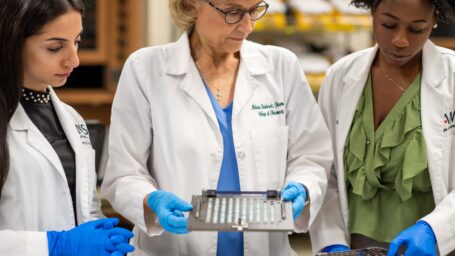 A professor holds medical equipment, looking to a student while another student looks to them. All three are wearing white coats and blue gloves