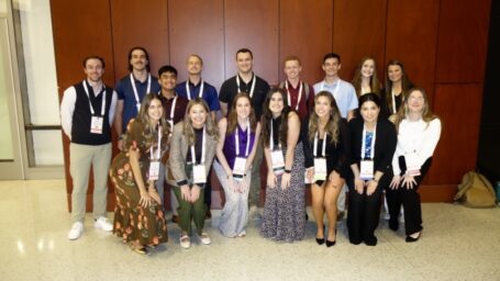 Group photo of 16 pharmacy students wearing event lanyards at a conference center